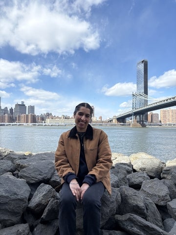 Harleen sitting on some large rocks beside a river with a bridge and city landscape in the back