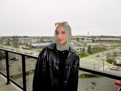 Ayesha stands on a balcony wearing a green scarf and a black jacket, with buildings and roads visible in the background.