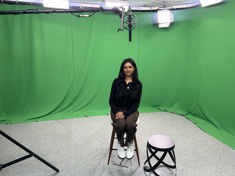 Rasana Yogarajan sitting on a stool in front of a green screen in a studio.