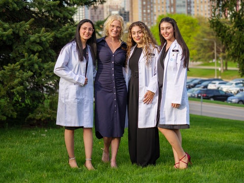 Four people stand together outdoors on a grassy lawn. Three women wearing white lab coats stand beside a woman in a navy dress in the center. Trees, a parking lot and campus buildings are visible in the background.