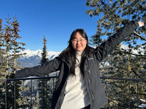 Hannah standing outdoors with their arms outstretched, smiling in front of snow-covered mountains and evergreen trees on a clear winter day.