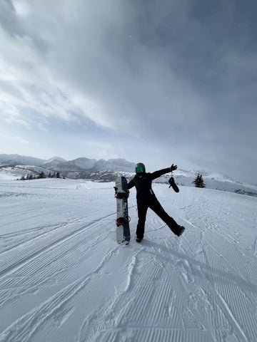 Hannah standing on a snowy ski slope with their skis upright beside her, posing against a backdrop of mountains and cloudy winter skies.