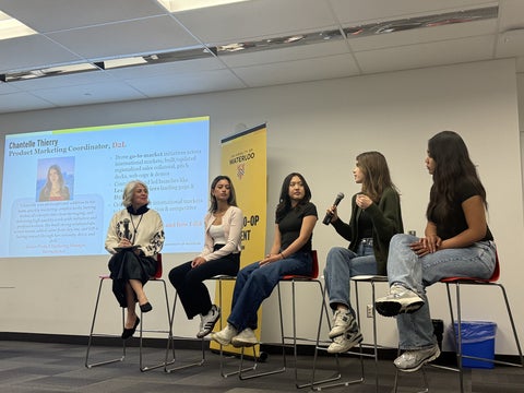: Five students sit on a panel at the front of a classroom, speaking into microphones during a discussion while a presentation slide is projected behind them.