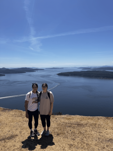 Harleen smiling with a friend standing on a mountain with scenery of water in the back