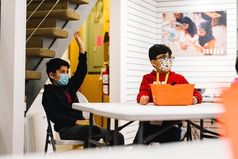 Two young students sitting at a table with one student raising their hand at LAUNCH Waterloo