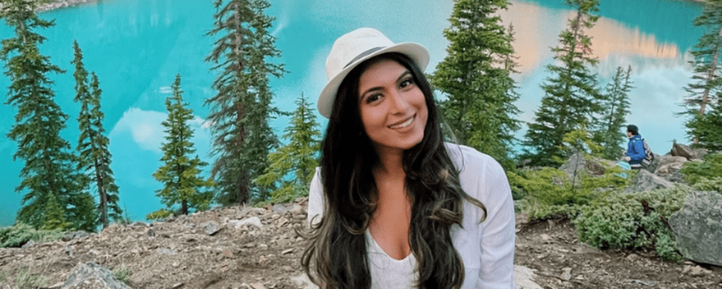 University of Waterloo Arts co-op student, Rashmi Jayaram, smiling outside, with pine trees and water in the background.