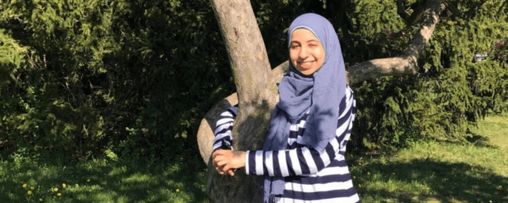 A photo of Fatima standing and smiling with her arms wrapped around a tree.