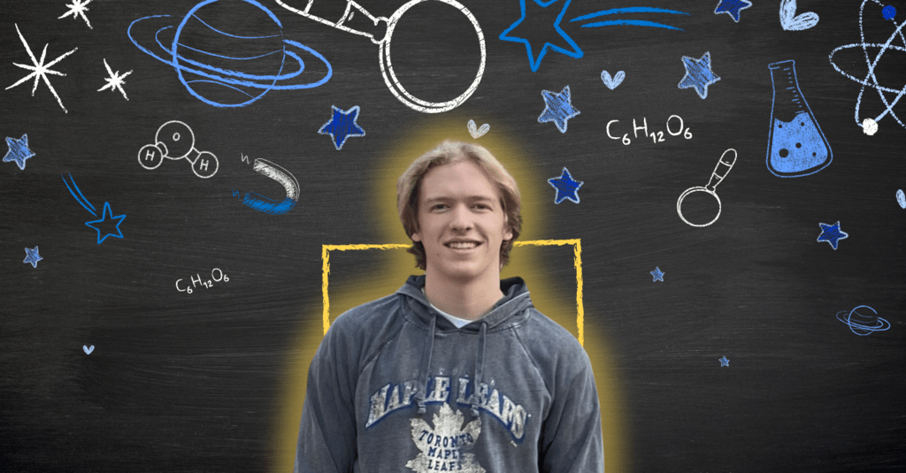 Luke smiling in front of a black board with blue and white science chalk drawings on it,