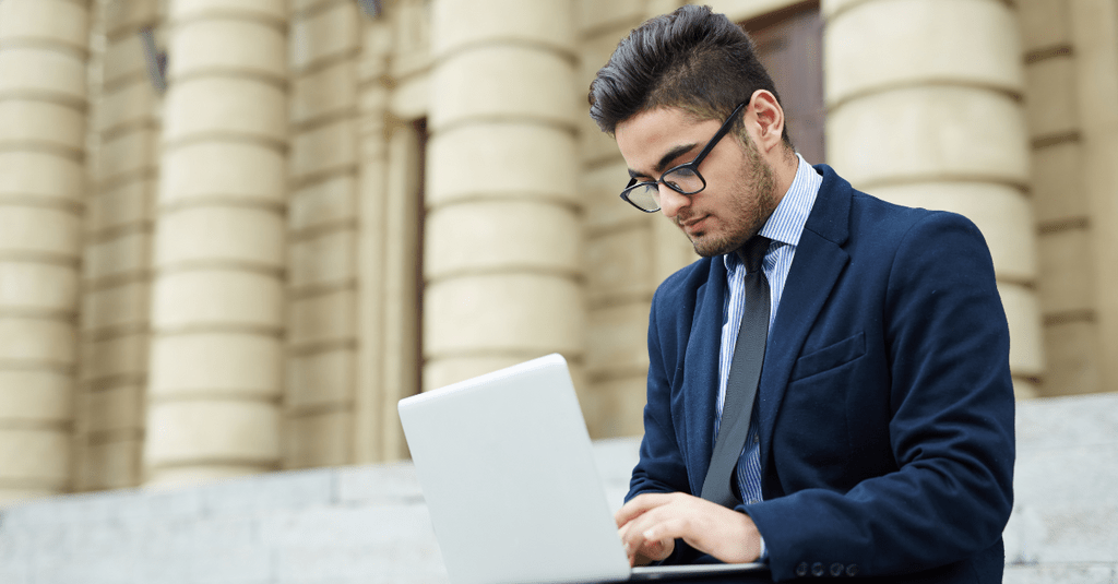 Male student working on his laptop sitting on the steps in front of a government building
