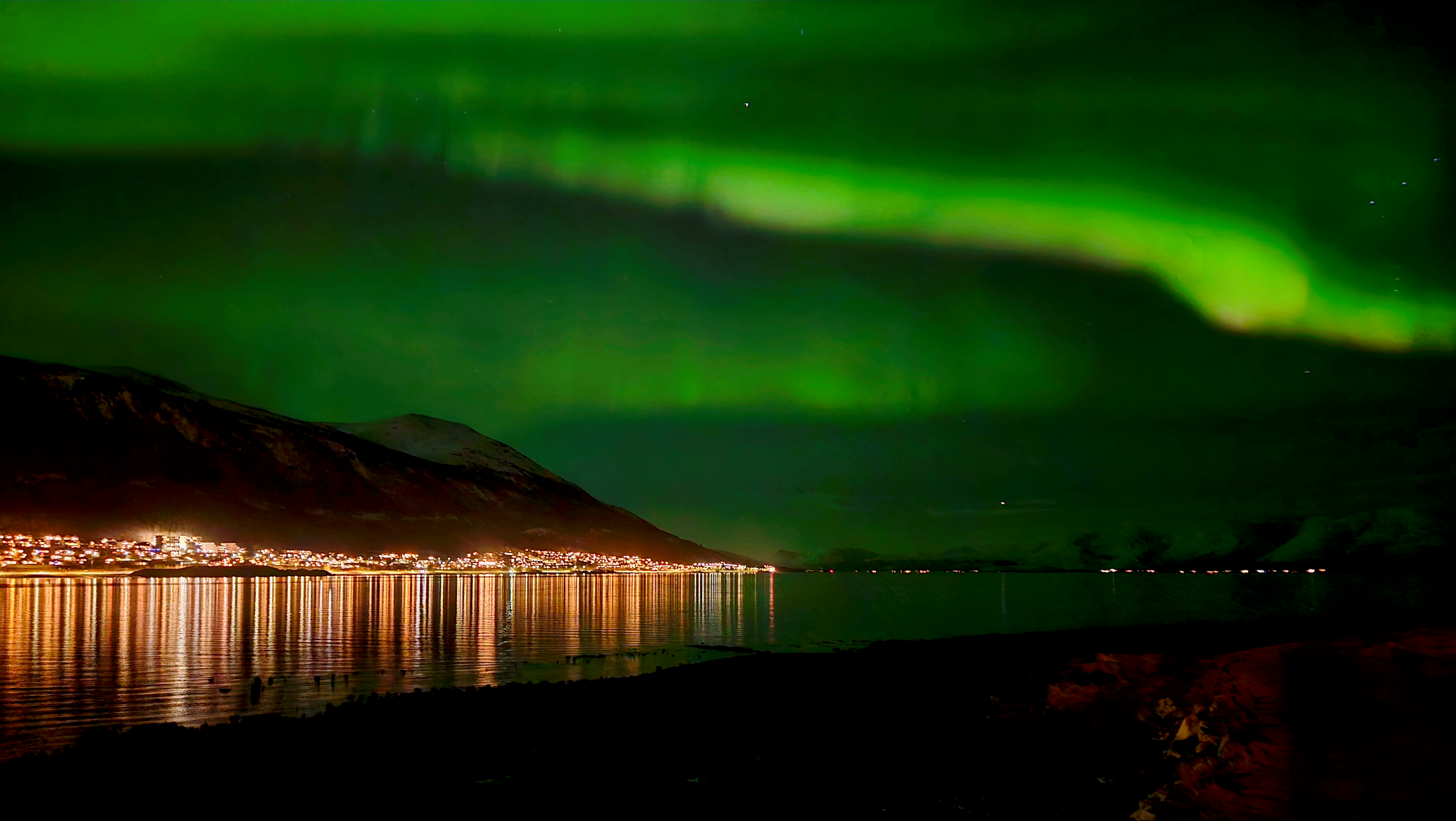 Green northern lights spread across a dark sky with a mountain range and the lights of town reflecting on black water.