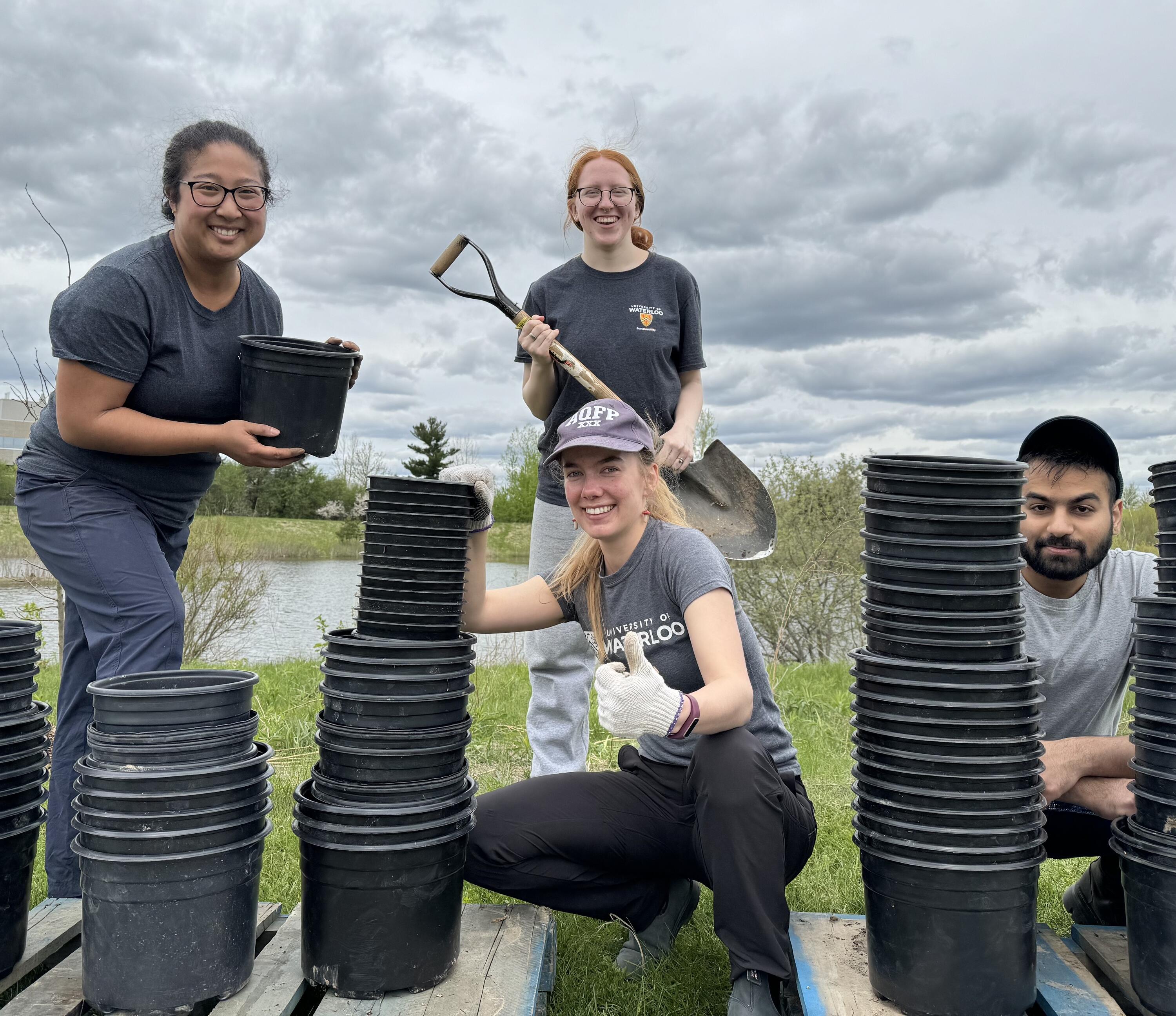 Alison Baird and three other students outside and smiling behind many empty pots. Alison is holding a shovel.