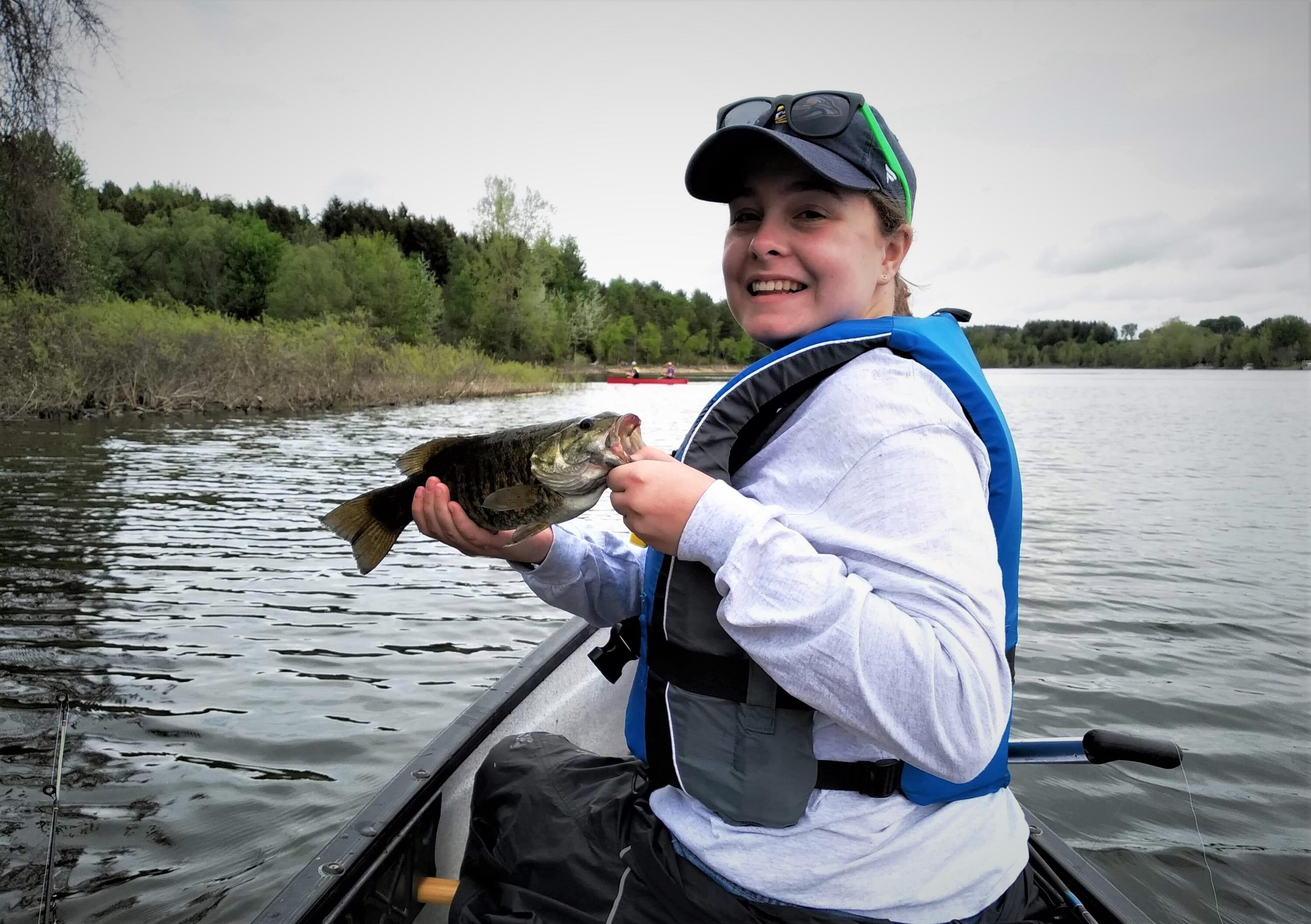 Photo of Daynica smiling while sitting on a canoe in the water and holding a fish