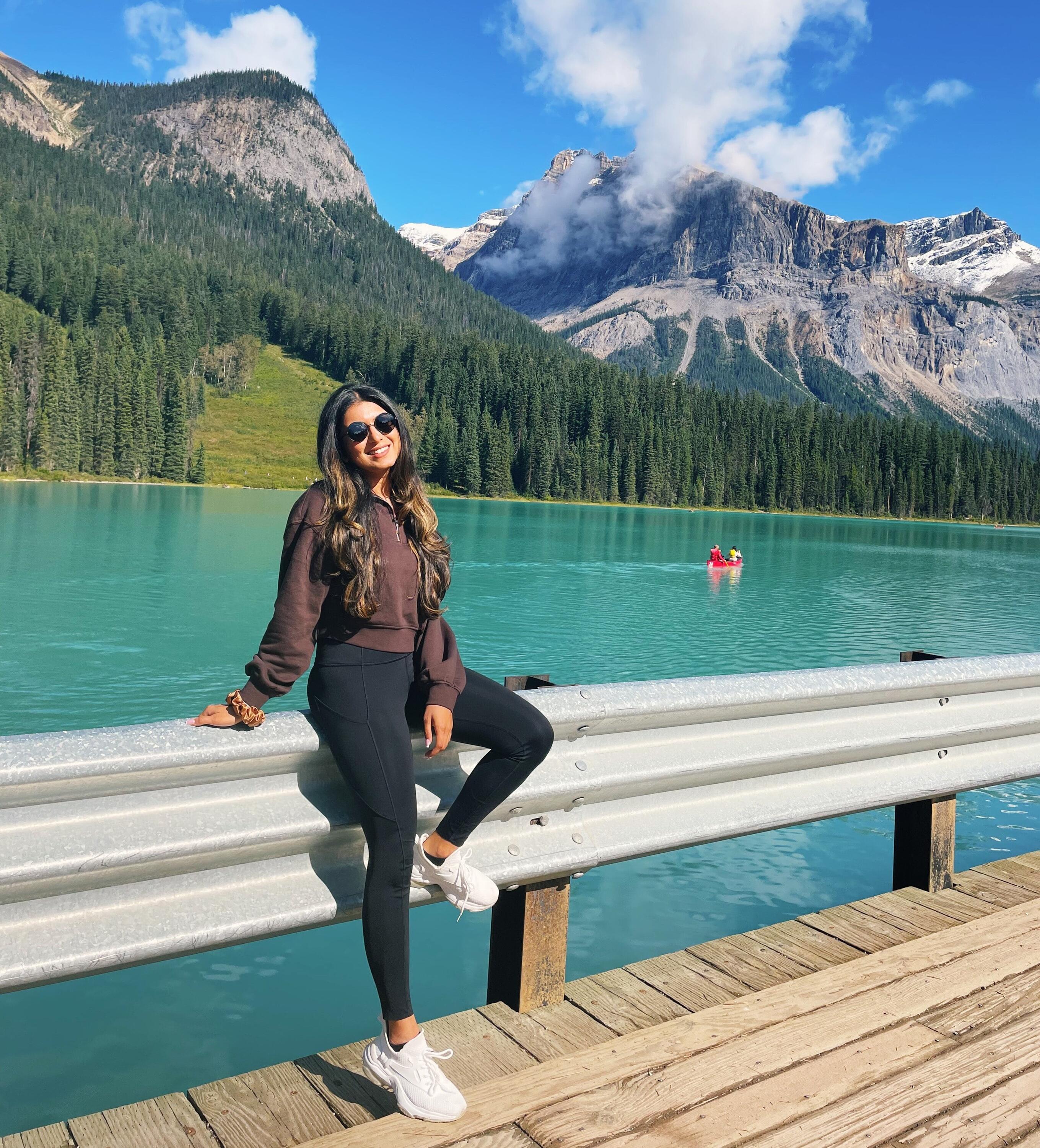 A photo of Rashmi smiling outside while leaning on a guardrail, with water, green trees, and mountains behind her.