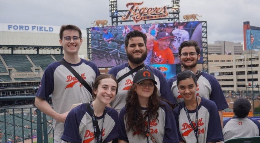 Taylor and 4 others wearing jerseys and in front of a Tiger's sign.