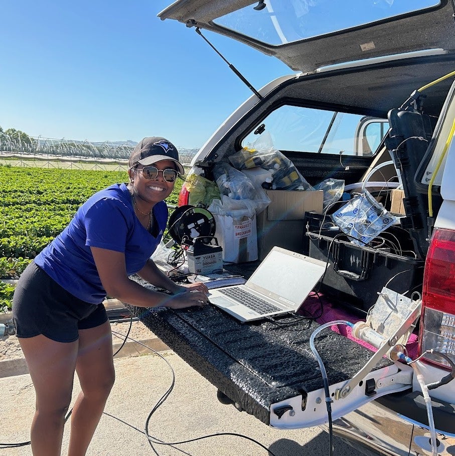 Thenuja Vittanachchi standing behind a car with an open trunk. The trunk contains various pieces of equipment and a laptop.