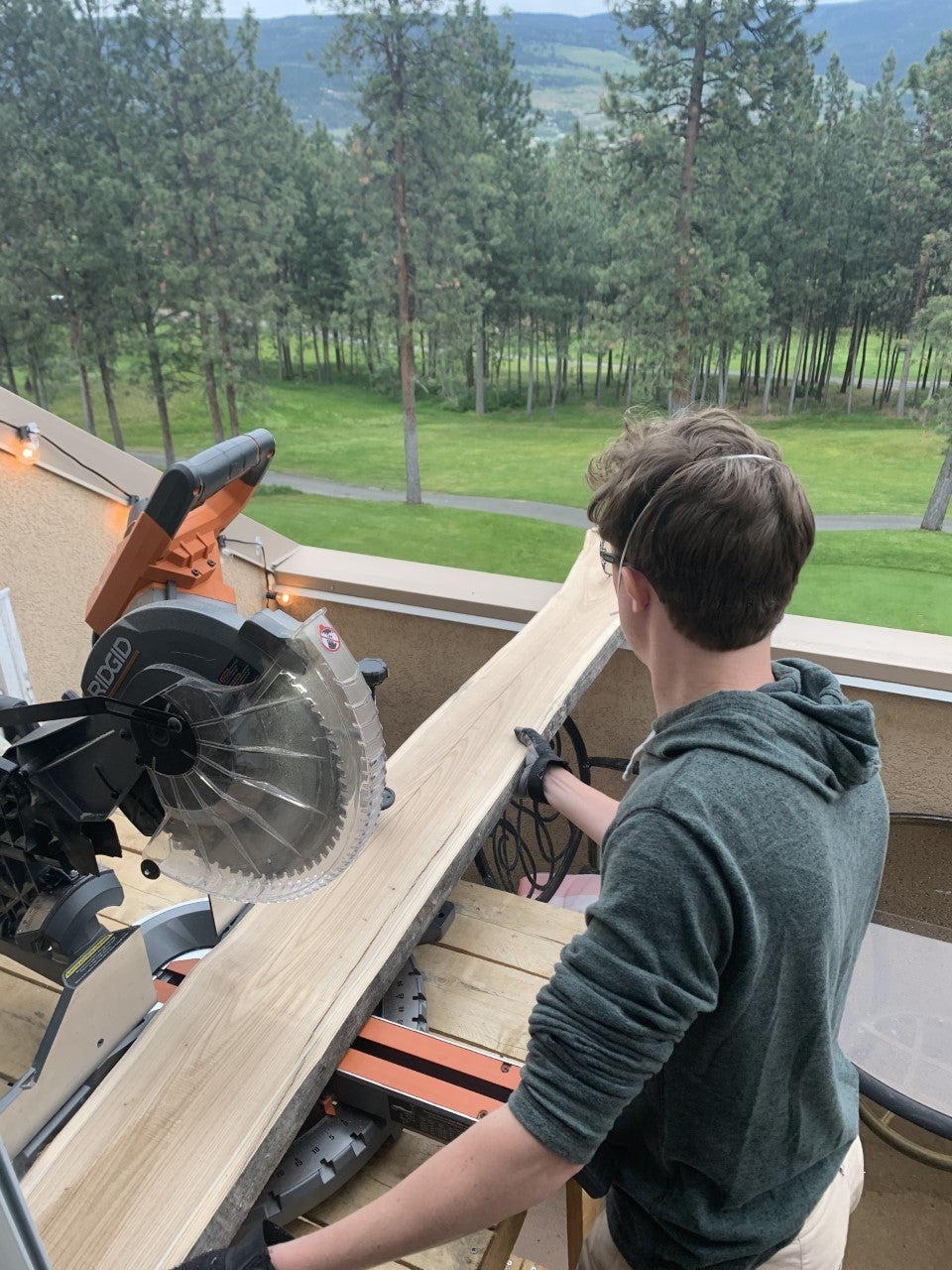 Photo of a man who is about to slice wood with a wood cutter machine, with trees and grass in the background.