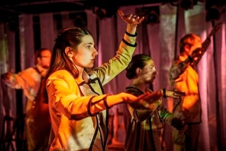 Performers in te middle of a slow dance routine surrounded by an orange and yellow lit set of poles that look like trees