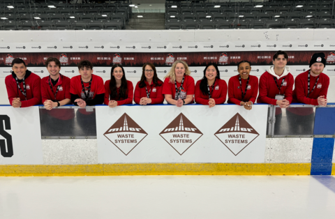 Event staff and volunteers wearing red shirts pose together along the rink boards at a hockey arena, smiling for a group photo during a tournament.