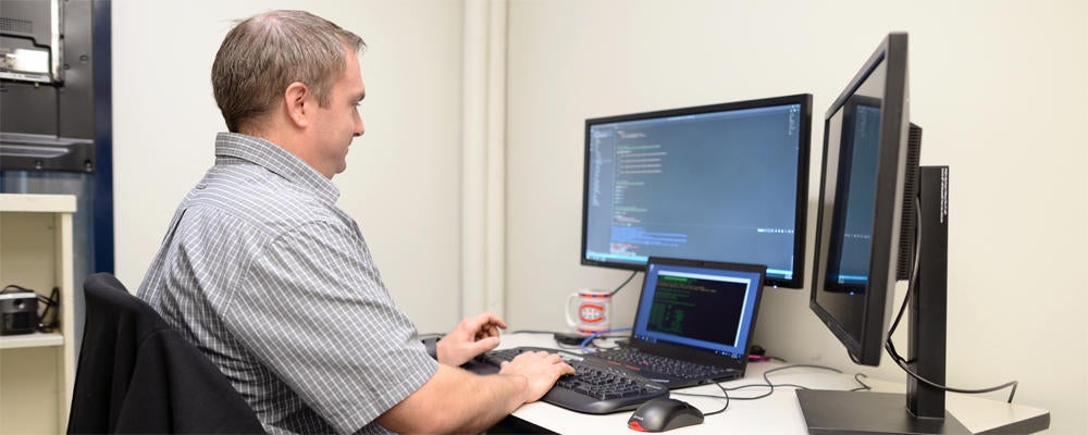 Staff member (Todd) works on some code at a workstation with three displays and a Montreal Canadiens mug.