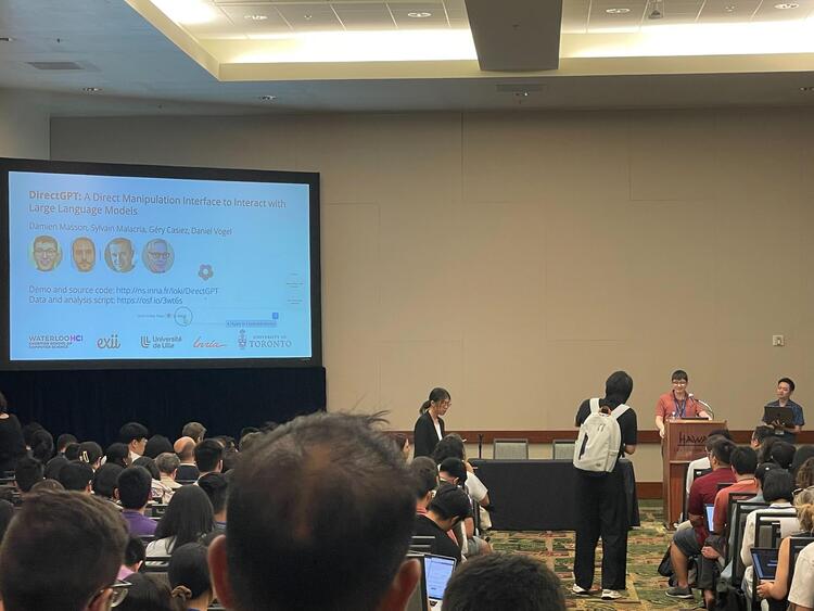 Photo of Damien in front of a podium, presenting their research to a large crowd. Background shows a slideshow with the paper's and authors names  