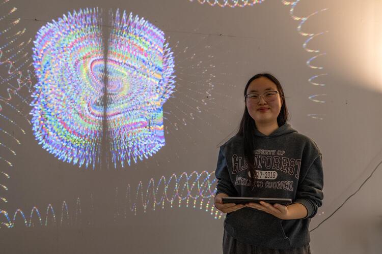 The artist standing in front of black screen. the middle is a drawing (shape is large oval) that has various mult-coloured waves inside. Surrounding the blob is random multi-coloured waves