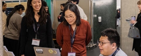 A group of people is gathered at a table with laptops. Two individuals stand, while one person is seated, looking at the screen. They are attending a research poster session.