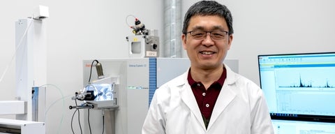 Person in a white lab coat standing in a laboratory with scientific equipment (mass spectronomy) and a computer screen.