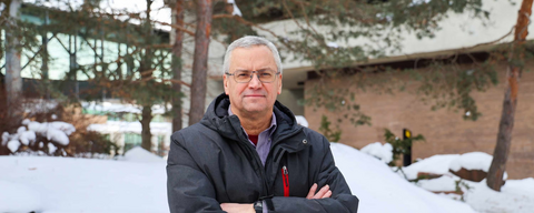 A man donning a black winter jacket posing in front of snow and brown building