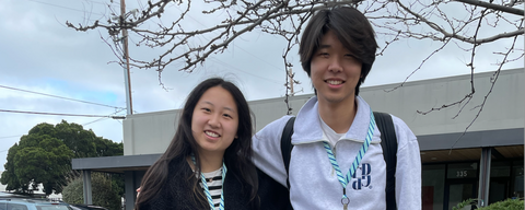 an asian girl in a striped black-and-white shirt and a black jacket standing next to an Asian man in a white hoodie. In the background you see a tree branch, a white building