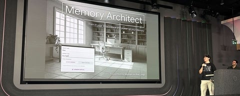 Stage presentation with a large screen titled “Memory Architect”; grayscale slide shows a room with shelves and a desk. Two presenters stand to the right, one speaking into a microphone.