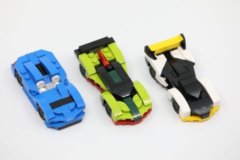 Three lego cars. left is a blue and black car with white seats, middle is a green-dominate black & red car, and last is a white and black car with yellow wings