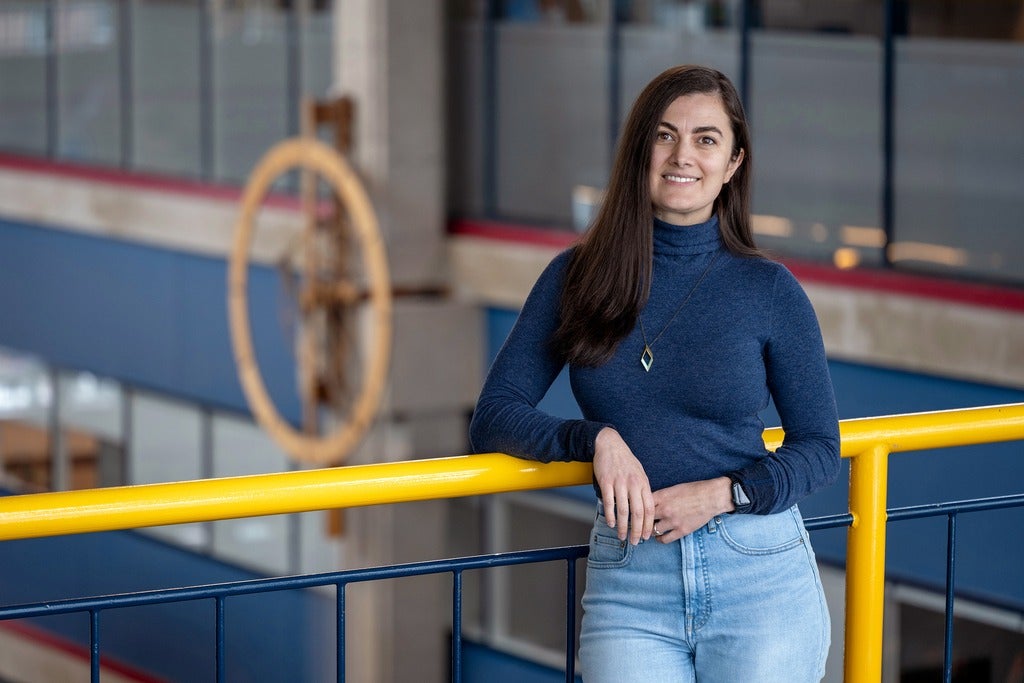 A brunette donning a dark blue long-sleeve sweater and diamond shaped necklace, leaning against a yellow railing. A large wooden clock is in the background, aganist blue, grey and red walls