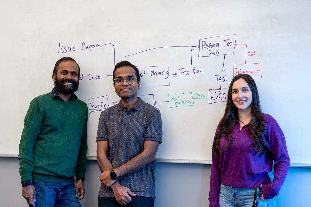 Three people (two men on the left and one woman on the right) stand in front of a whiteboard filled with a flowchart; left wears a green sweater, middle a gray dotted shirt, right a purple top.
