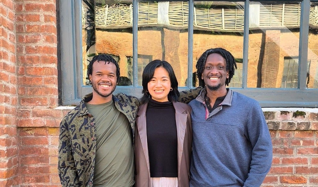 Photo of three co-founders posing against a red-brown brick wall with a large blue window. Left the co-founder is wearing a camo jacket with a muddy green shirt. The middle co-founder is wearing a black shirt and a brown leather jacket. The right co-founder is wearing a blue sweater