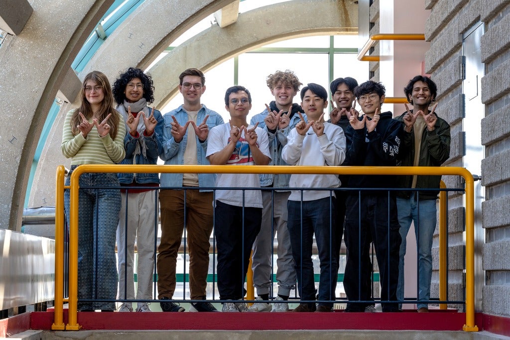 9 waterloo students standing behind a yellow bar and doing the "W" sign with their hands