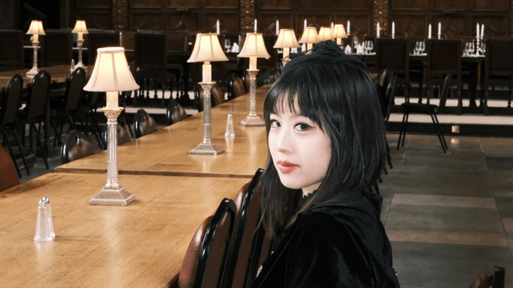 A person in a black velvet dress and gloves sits at a long wooden table in a historic dining hall with decorative wall panels and lamps. A large painting hangs on the wall behind them.