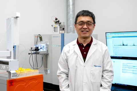 Person in a white lab coat standing in a laboratory with scientific equipment (mass spectrometry) and a computer screen.