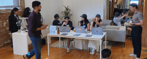 A group of people at a table with laptops and drinks in a casual office space, while another person walks by.