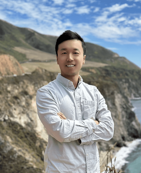 An Asian man in a white collar up shirt posing in front of mountains. in the right bottom corner you can see glimpses of a lake/sea