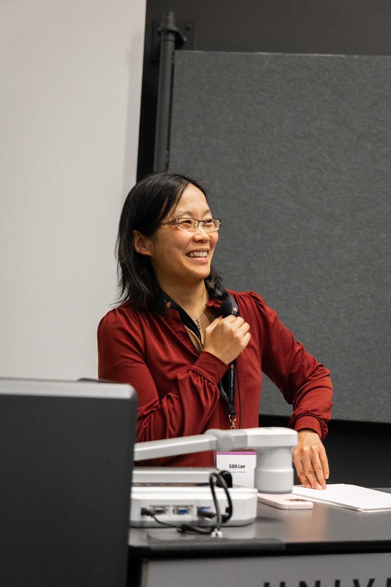 An Asian woman holding a mic and chuckling in front of a desk. She is wearing a red long sleeve blouse