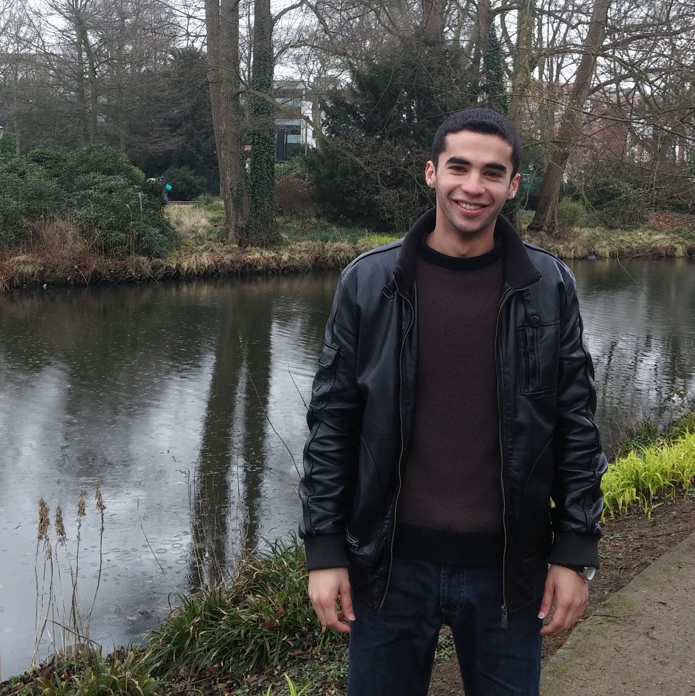 Ahmed Alquraan: a South Asian man donning a dark sweater and jacket posing in front of a creek