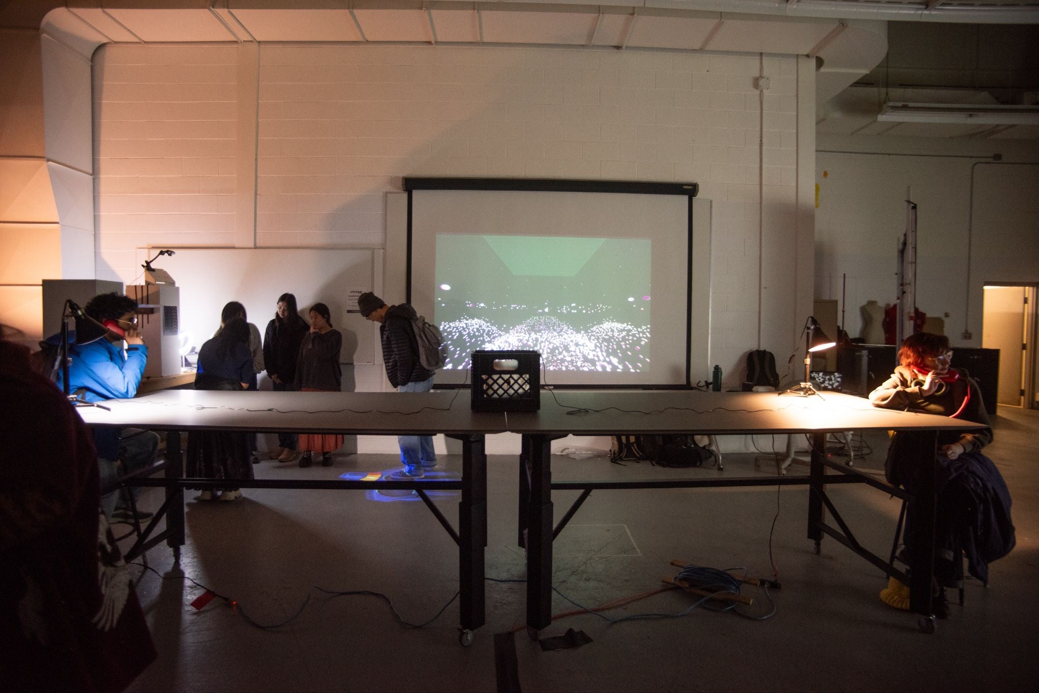 2 people sitting on opposite ends of a long table. They are talking on retro-like phones. The phone's wires are connected to a laptop that’s placed in a black crate in the middle of the table. The background is very dimly lit