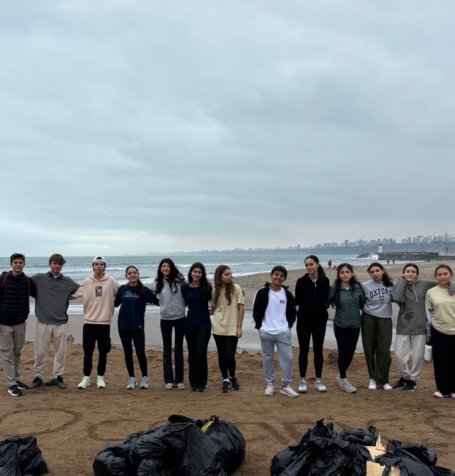 A group of people (wearing hoodies and long sleeve shirts) standing side-by-side on a beach. In front of them are trash bags 
