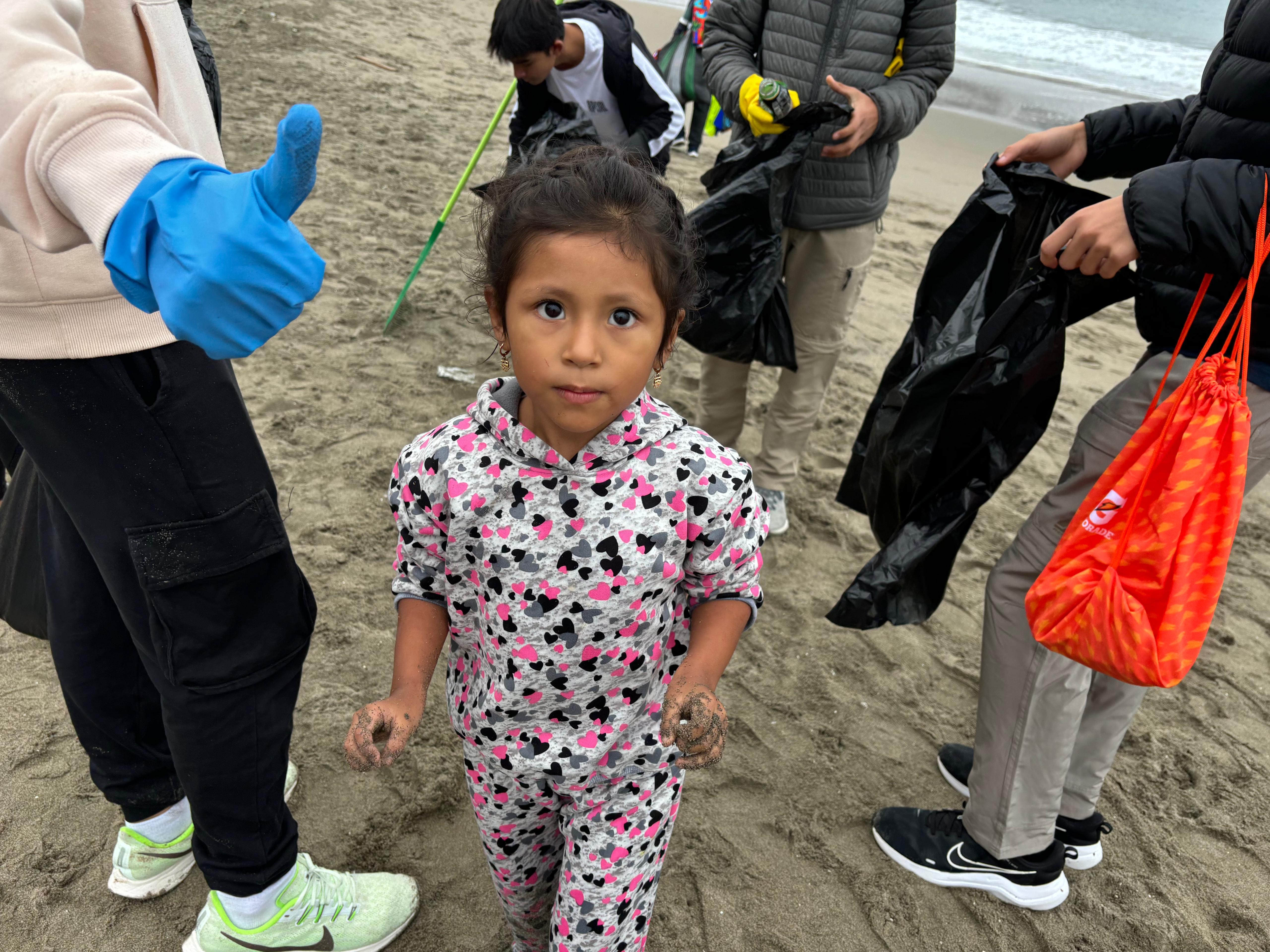 A close-up of the people cleaning a beach (holding trash bags). You can see someone's hand donning a blue cleaning glove and giving a thumb-ups (face not visible). In the middle is a young girl facing the camera