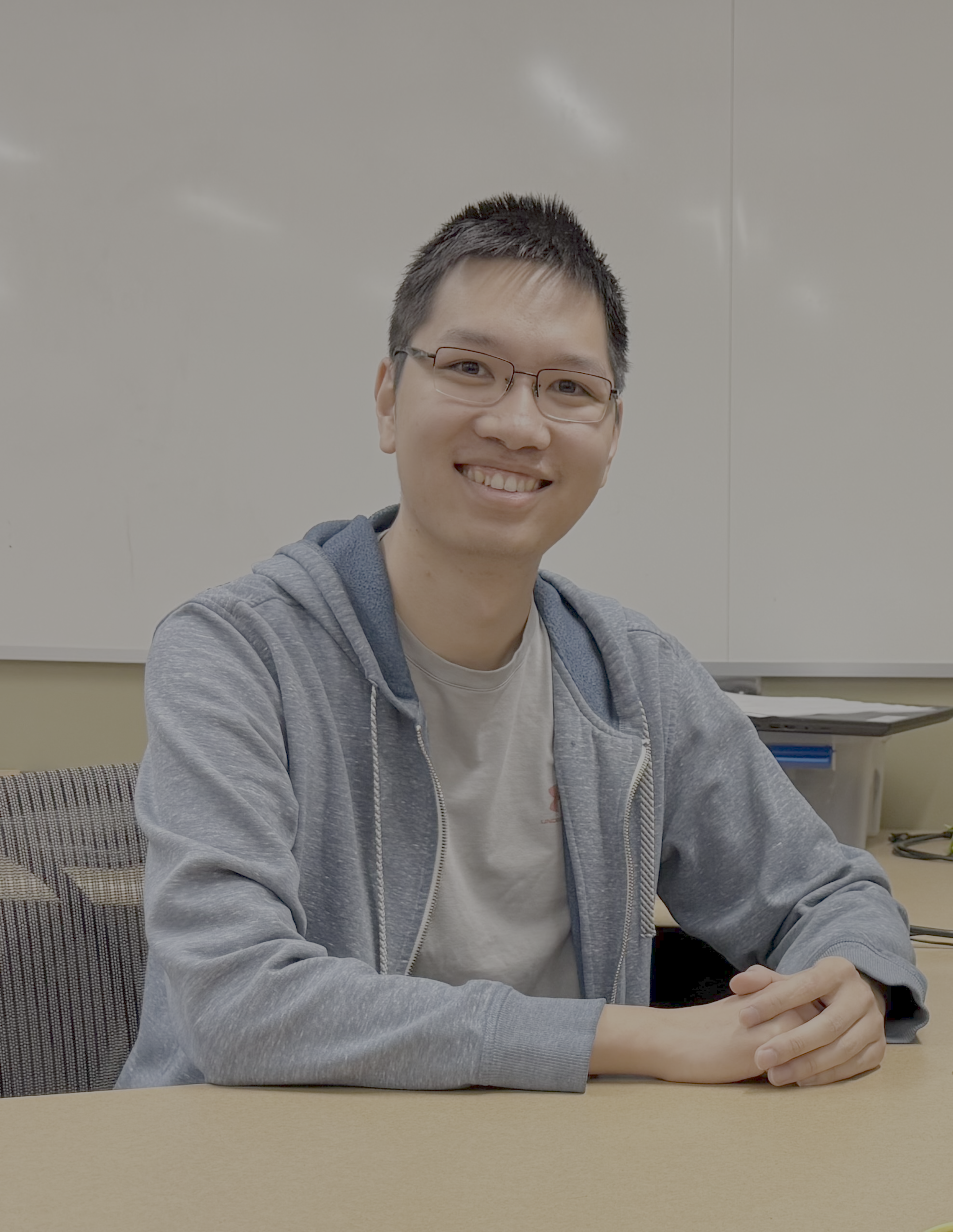 An Asian man wearing a blue hoodie and grey shirt sitting down in front of a whiteboard. His hands are clasped on the table and