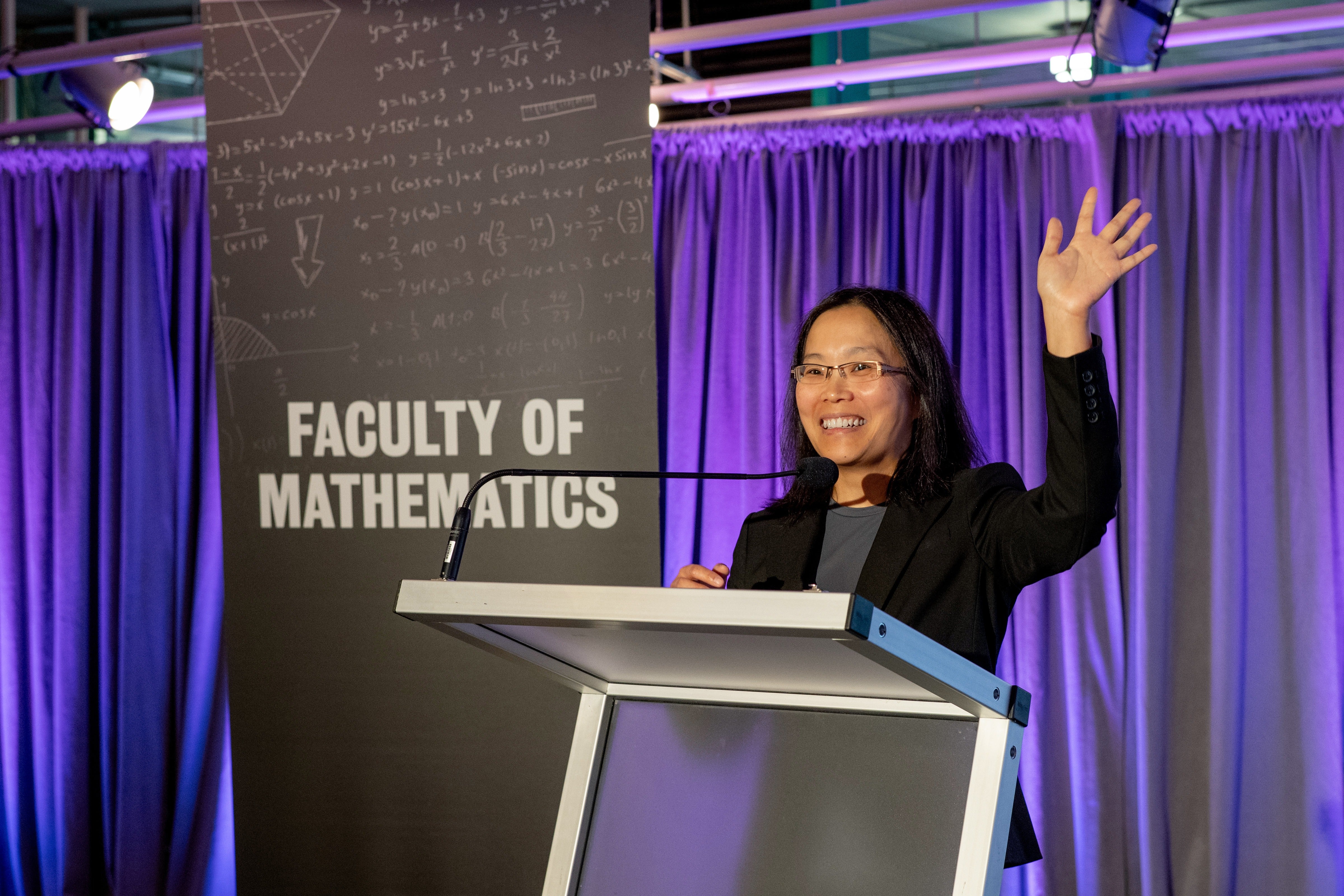 Edith Law wearing a black blazer and grey shirt and waving to the crowd (not shown) the backdrop includes purple curtains and a tall black sign reading "faculy of Math" with math symbols