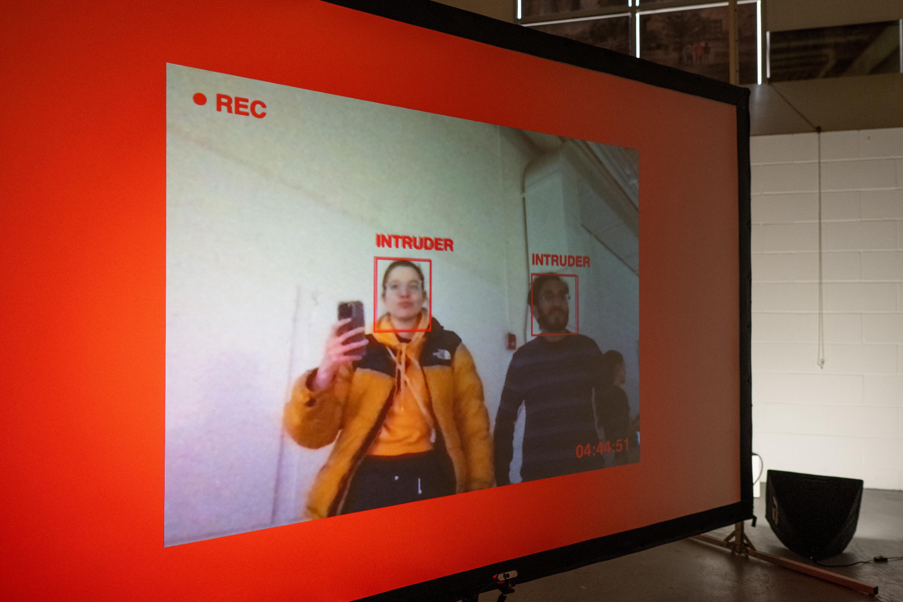  background is red. inside if a smaller recentage mimicing a video feed (you can see 2 people). Their faces are marked with a red rectange, with the word "intruder" and the left top corneer is a REC sign, bottom right corner is a timestamp