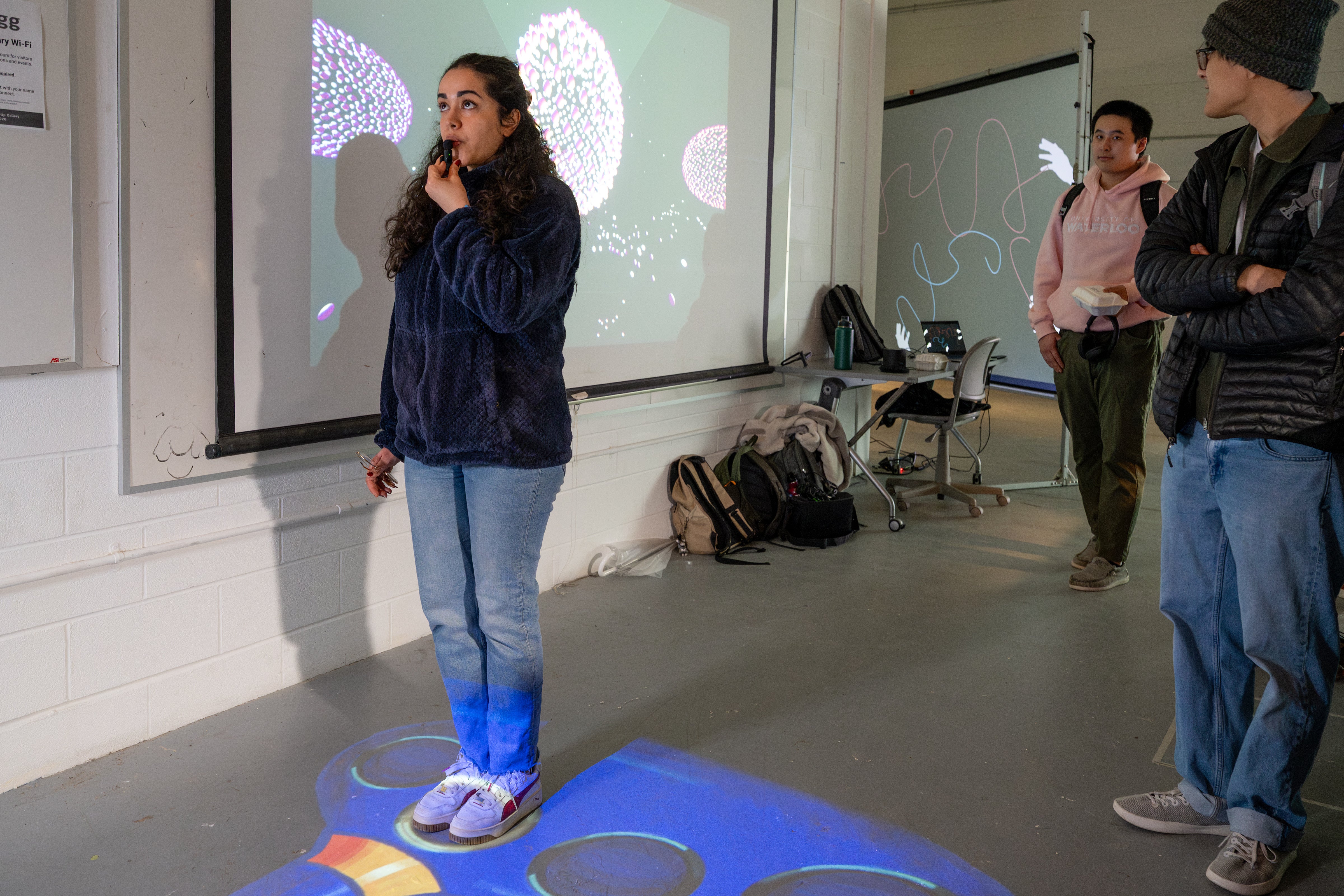 A woman holding and blowing into a small microphone. She's standing on the "Floor Carina" projection, which shows a blue-coloured instrument with holes. The colouring and shape kinda looks like the Genie's bottle from Aladdin.