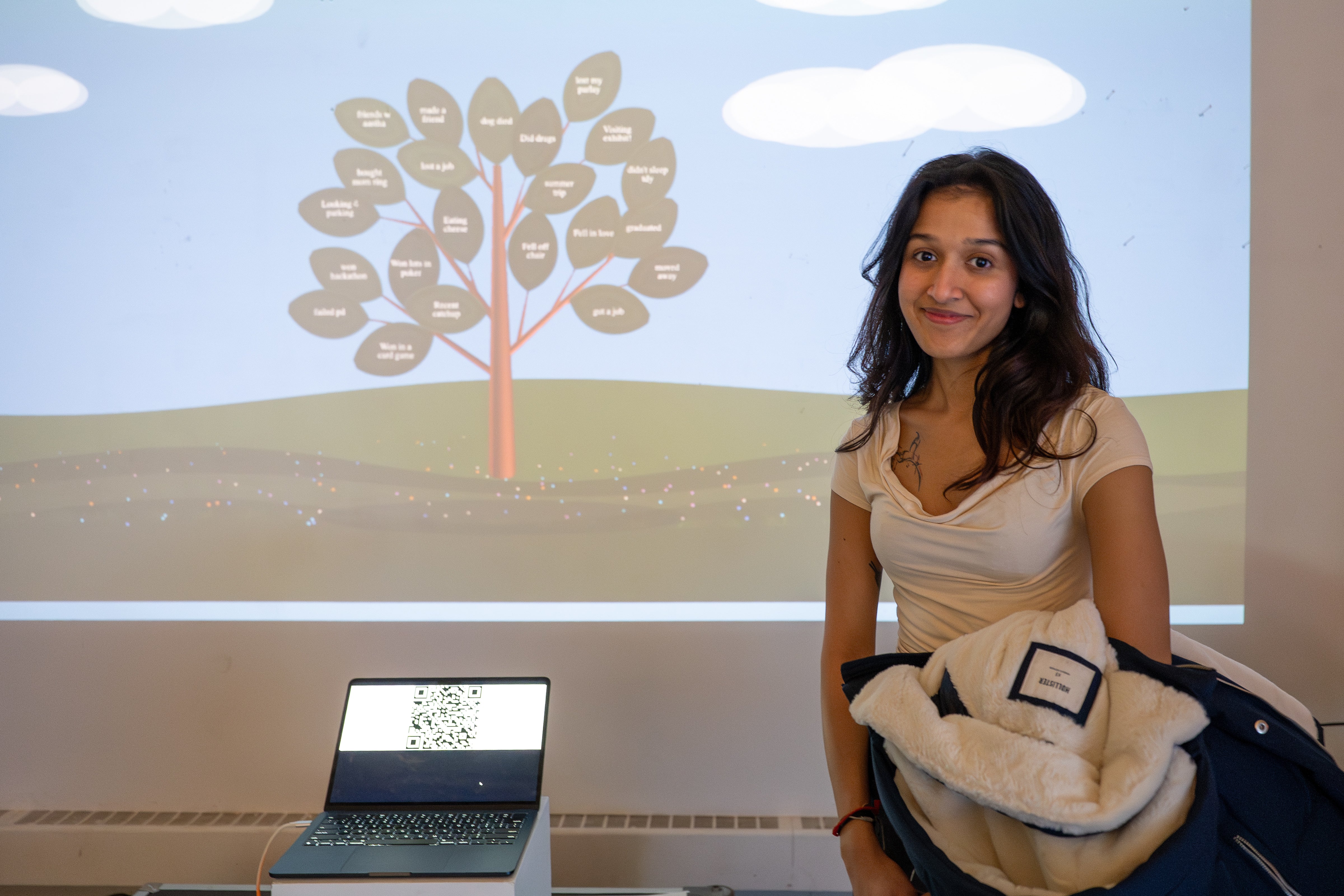 A South Asian woman posing in front of a projection of her artwork, Memory Tree. It depicts a growing tree with leaves that present user-submitted memories (ex. "Ate Mango" "Got Fired")