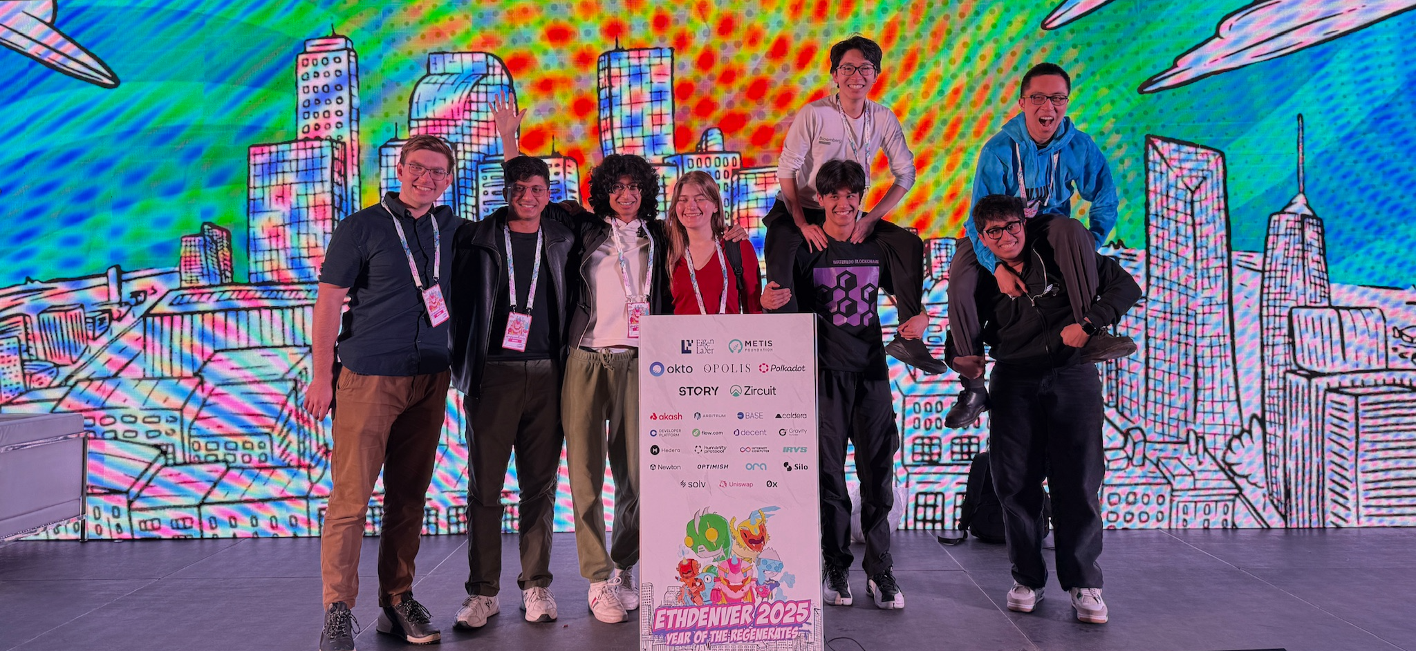 A group of eight people poses in front of a colorful, illustrated cityscape backdrop, holding a sign with event details.